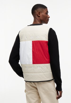 Άνδρας φοράει γιλέκο με χρώματα μπεζ, κόκκινο και λευκό, με την επιγραφή "Tommy Jeans" στην πλάτη, συνδυασμένο με μαύρο πουλόβερ και μπεζ παντελόνι.