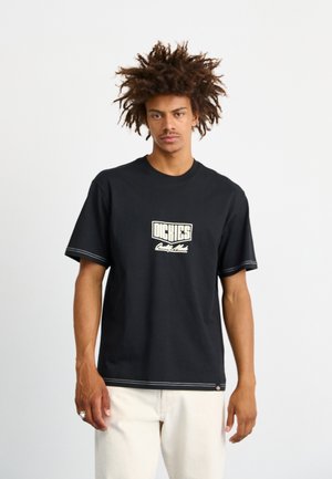 T-shirt nera in cotone con maniche corte, caratterizzata da cuciture bianche e un logo al centro. Vestibilità semplice e rilassata, abbinata a pantaloni chiari.