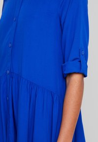 Robe boutonnée bleue à manches courtes retroussées et taille froncée pour une silhouette fluide, fabriquée en tissu léger.