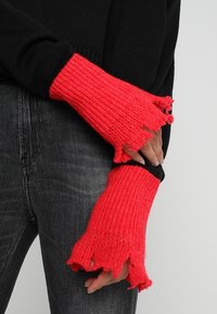Rote gestrickte fingerlose Handschuhe mit geripptem Muster und ausgefransten Kanten, kombiniert mit einem schwarzen Pullover und grauen Jeans.