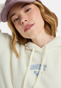 Crème fleece hoodie met een blauwe "ROXY" logo, koordjes en een relaxte pasvorm, gecombineerd met een lichtpaarse corduroy pet.