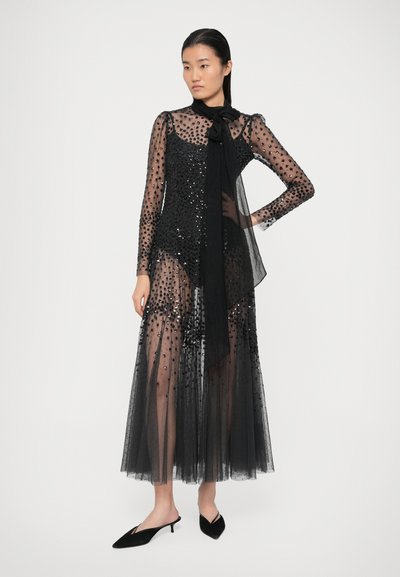 Nais seiseb seljas läbipaistva musta pika kleidi, millel on täpid, sädelused, kaelabow ja mustad terava ninaga väikese kontsaga kingad.