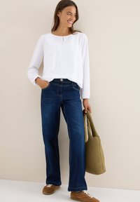 Blouse blanche à manches longues avec un détail en trou de serrure, associée à un jean taille haute bleu foncé et des baskets brunes, tenant un sac vert.