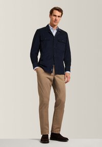 Giacca blu navy con bottoni, dotata di due tasche sul petto, abbinata a pantaloni beige e stivaletti alla caviglia marrone scuro. L'outfit presenta un design con colletto.