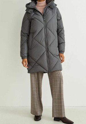 Personne portant un long manteau matelassé gris avec une capuche, un col roulé rose, un pantalon large à carreaux et des bottines marron foncé.