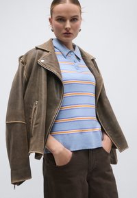 Καφέ δερμάτινο μπουφάν με ασημί φερμουάρ, ανοιχτό μπλε πόλο με ρίγες σε κίτρινες και μπλε αποχρώσεις, σκούρο καφέ παντελόνι.