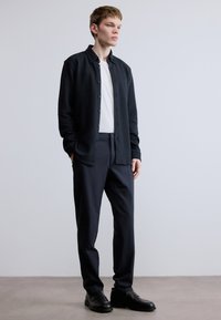 Tmavá košeľa na zapínanie z mäkkej látky, kombinovaná s bielym tričkom s okrúhlym výstrihom a strihanými tmavými nohavicami. Obuv tvoria čierne kožené topánky.