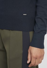 Maglione girocollo blu navy con polsini e orlo a coste. Include un'etichetta in metallo con il logo. Abbinato a pantaloni verde oliva con strisce laterali scure.