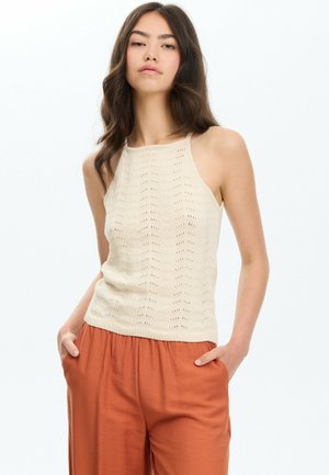 Mujer joven con cabello largo y oscuro lleva una camiseta de punto sin mangas color crema y pantalones holgados de color óxido con las manos en los bolsillos.