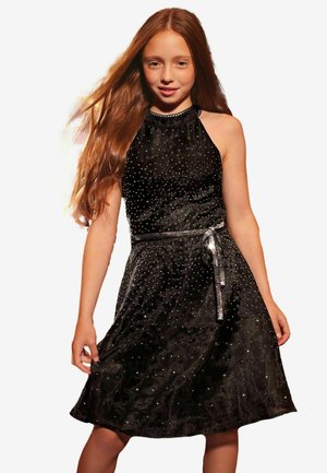 Niña con el pelo largo y rojo que lleva un vestido de terciopelo negro sin mangas con pequeños adornos brillantes y un cinturón brillante atado a la cintura.