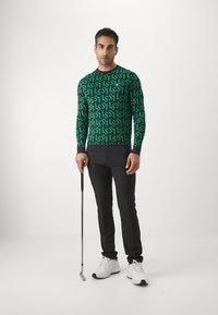 Maglione verde a maniche lunghe con motivo geometrico nero, abbinato a pantaloni neri e scarpe da ginnastica bianche; tiene in mano una mazza da golf.
