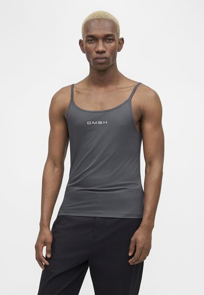 Νέος άνδρας με κοντά ξανθά μαλλιά, που φοράει εφαρμοστό σκούρο γκρι αμάνικο μπλουζάκι με το κείμενο "GMBH" και χαλαρό μαύρο παντελόνι, σε απλό φόντο.