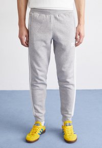 Grå sweatpants med vita sidostråkar, elastisk midja och en mjuk textur, som bärs med gula sneakers med blå detaljer.