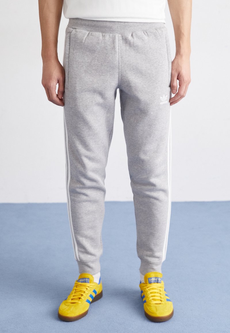 Grå sweatpants med vita sidostråkar, elastisk midja och en mjuk textur, som bärs med gula sneakers med blå detaljer.
