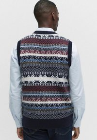 Mörkblå stickad väst med en flerfärgad geometrisk mönstring, ribbad halsringning och enfärgad mörkblå nederkant, bärs över en ljusblå skjorta.