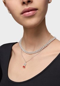 Collar multihebra de plata con piedras brillantes y un colgante que presenta una gema roja y clara. Lleva puesto sobre una camiseta negra.