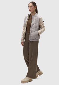 Vesta matlasată de culoare gri deschis cu mâneci bej, combinată cu pantaloni largi maroni și cizme bej. Materiale texturate și design ajustat.