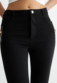 Melnas pieguļošas bikses ar metāla pogu, priekšējo rāvējslēdzēju un dekoratīvām sānu kabatām. Gluda tekstūra ar elastīgu materiālu komfortam.