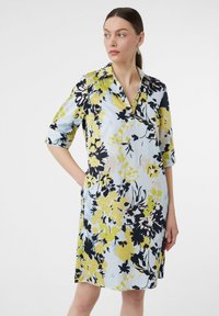 Blumenkleid aus leichtblauem Stoff mit großen gelben und schwarzen Blumendrucken, kurzen umgeschlagenen Ärmeln und einem kragenförmigen Halsausschnitt.