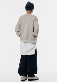 Felpa beige oversize con spalle abbassate, indossata sopra una maglietta bianca. Abbinata a pantaloni neri larghi e stivali grigi. Berretto a coste scuro.