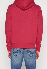 Czerwona bluza z kapturem o długich rękawach, z prążkowanymi mankietami i dołem, zestawiona z niebieskimi dżinsami z charakterystycznym przeszyciem na tylnych kieszeniach.