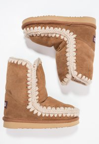 Des bottes en suede marron présentent une accent crème tressée le long des coutures, un intérieur doux et une semelle en caoutchouc plate avec un léger talon.