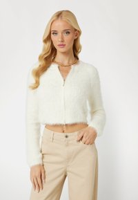 Witte, luchtige, cropped cardigan met lange mouwen en een ritssluiting aan de voorkant. Draagt over beige broeken, met een zachte textuur en een subtiele glans.