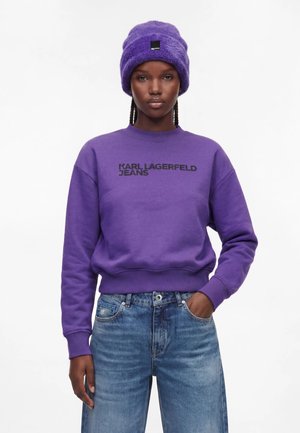 Vrouw draagt een paarse Karl Lagerfeld Jeans trui, paarse pluizige muts en blauwe spijkerbroek, met één hand in de zak tegen een witte achtergrond.