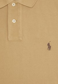 Beige textured polo triko se dvěma bílými knoflíky a malým tmavohnědým vyšitým logem hráče polo na hrudi.