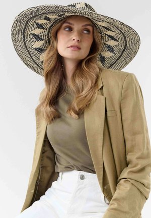 Woman with long wavy hair wearing a wide-brimmed patterned straw hat, olive green top, beige blazer, and white pants looking sideways.