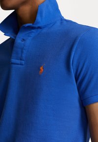 Homme portant un polo bleu vif avec col relevé et petit logo brodé orange de joueur de polo sur la poitrine.