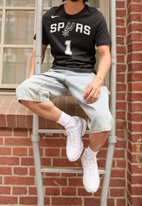 Czarny t-shirt z krótkim rękawem z napisem "SPURS" i numerem "1", jasnoniebieskie szorty dżinsowe, białe sneakersy i białe skarpetki do kostek. Przemysłowe tło.