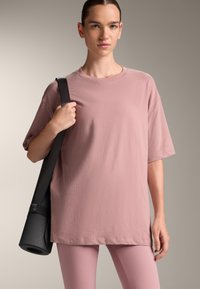 Oversized pink cotton T-shirt with short sleeves, paired with light pink leggings. A black yoga mat is slung over the shoulder.
