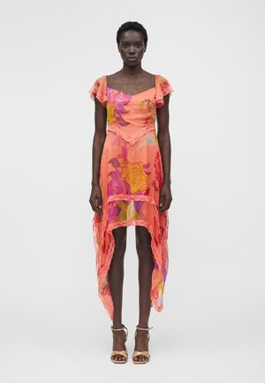 Kobieta ubrana w prześwitującą koralową sukienkę z wielokolorowymi wzorami kwiatowymi, koronkowym wykończeniem, krótkimi, falującymi rękawami i asymetrycznym dołem, w połączeniu z beżowymi szpilkami.