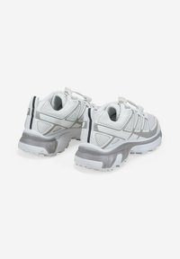 Par de zapatillas deportivas blancas y grises con paneles de malla, suelas gruesas y cordones elásticos atados, mostradas desde la parte trasera sobre un fondo blanco.