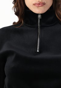 Svart fleece zip-up topp med høy krage, med en metallglidelås og merkevaremaskinvare, som viser en jevn tekstur og avslappet passform.