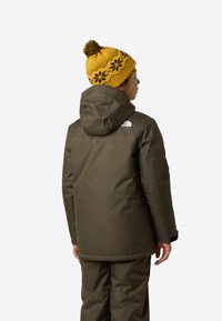 Brun isolert jakke med hette og The North Face-logo, kombinert med gul strikkelue med svarte blomsteraktige mønstre og dusk.