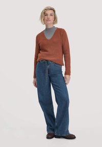 Rostfarbener, gestrickter Pullover mit V-Ausschnitt und geripptem Muster, kombiniert mit lockeren blauen Jeans und dunkelbraunen Schuhen.
