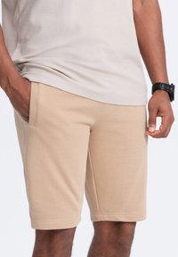 Shorts in cotone beige con una cintura elastica, tasche laterali e una texture liscia. Indossati con una maglietta beige chiaro e un orologio nero.