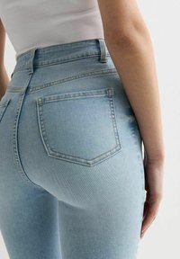 Detailní záběr na osobu v světle modrých džínách s vysokým pasem a bílém ribbovaném topu bez rukávů, zobrazení zadní kapsy a paže.