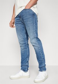 Világoskék, slim fit farmernadrág egyetlen érmepénztárcával és enyhe kopásokkal. Fehér magas szárú sportcipőkkel párosítva.