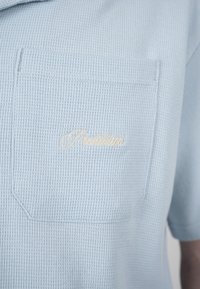 Chemise en tissu gaufré bleu clair avec une poche poitrine avant comportant un petit texte brodé en cursive avec un fil clair.