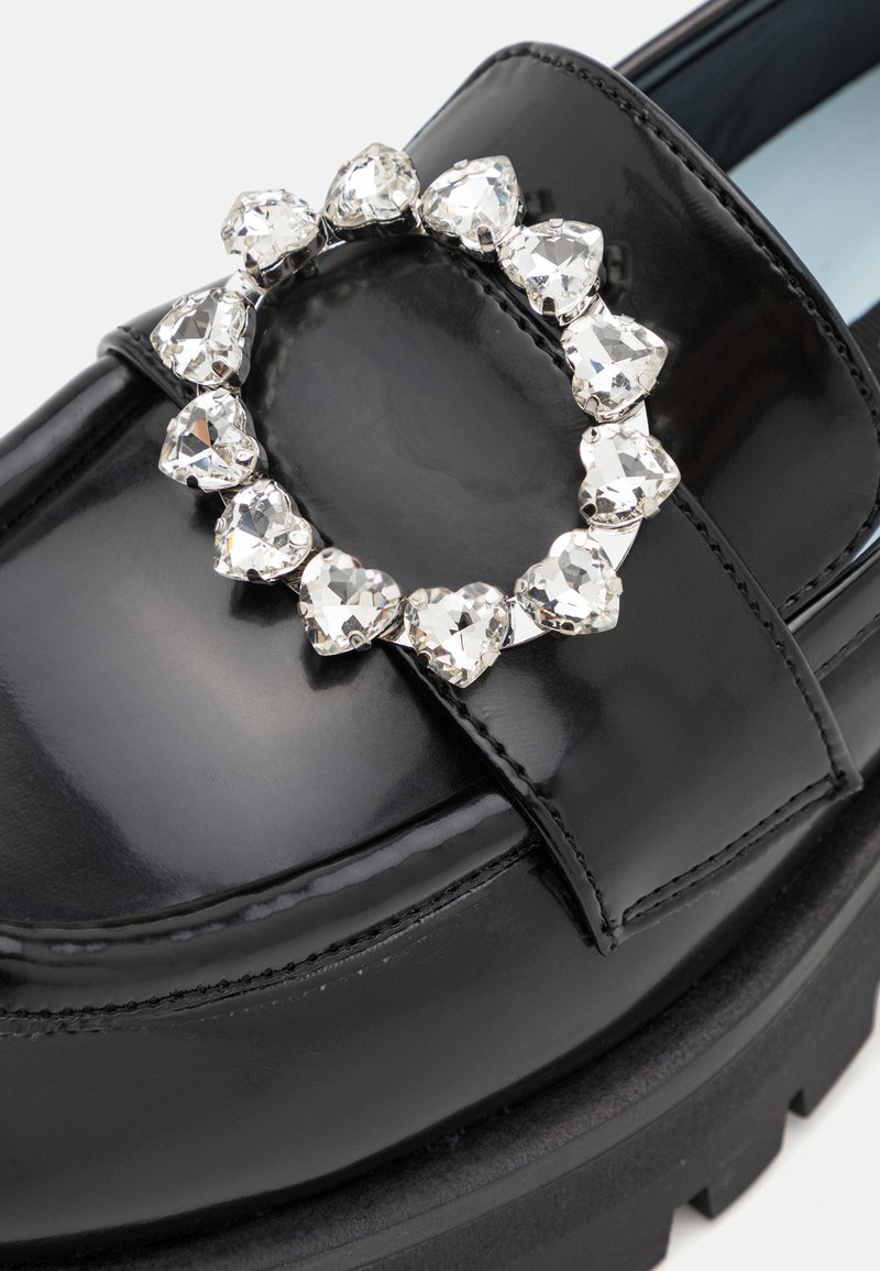 Mocassino in pelle verniciata nera con superficie testurizzata e suola spessa nera. Presenta un embellishment a forma di cuore in strass argentati sulla parte anteriore.
