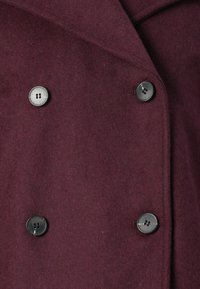 Dobbeltbrystet borst coat i mahogni med fire sorte knapper arrangeret i to lodrette rækker.
