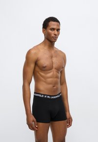 Juodi bokseriniai šortai iš lygaus audinio, su plačiu juosmens diržu, kuriame baltais raidėmis parašytas „KARL LAGERFELD“ logotipas. Aptempto dizaino, minimalus detalumas.