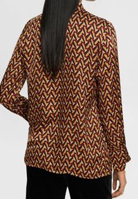 Femme avec de longs cheveux noirs portant une chemise à manches longues à motifs géométriques en rouge, moutarde, noir et beige, se tenant avec une main sur la hanche.