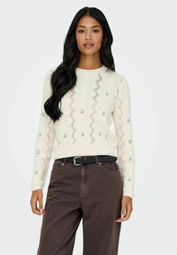 Pull en maille crème avec un motif en zigzag, orné de broderies florales bleues sur les manches. Assorti avec un pantalon taille haute marron et une ceinture noire.