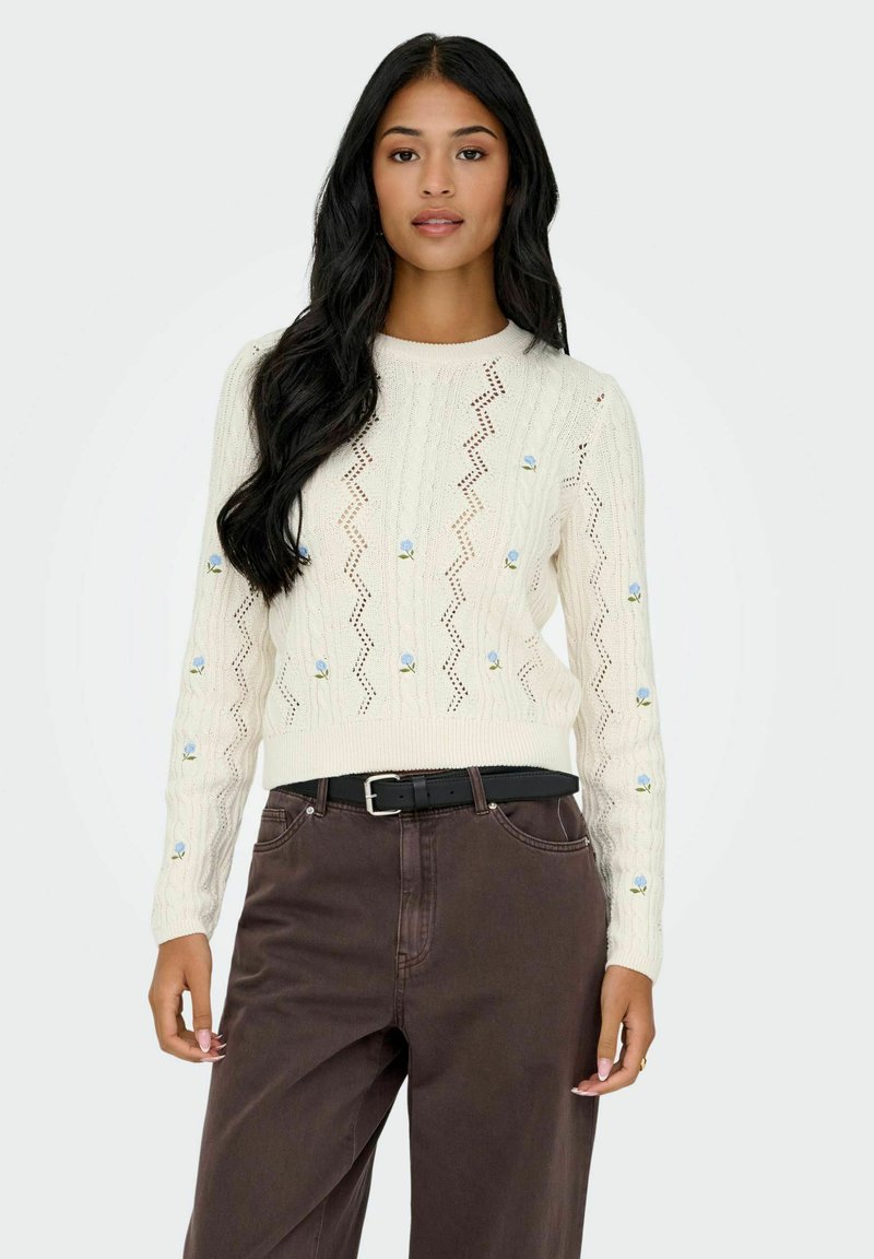 Pull en maille crème avec un motif en zigzag, orné de broderies florales bleues sur les manches. Assorti avec un pantalon taille haute marron et une ceinture noire.