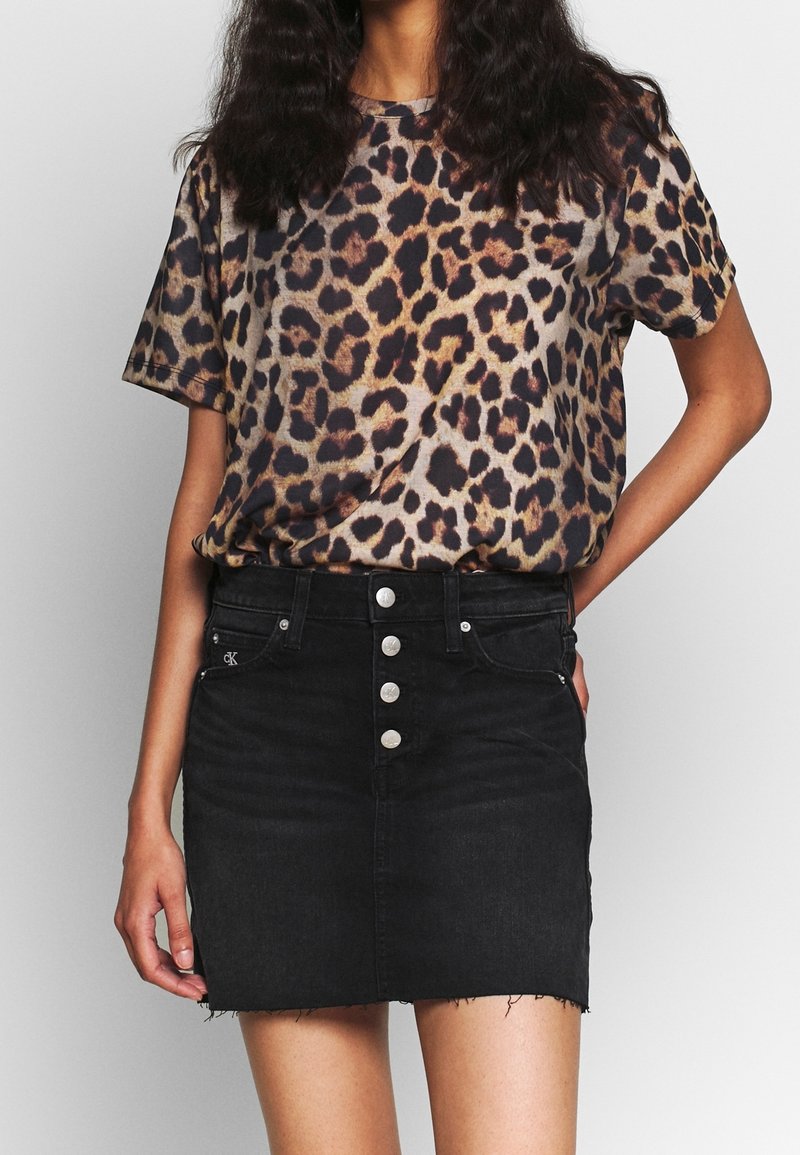 Kvinna klädd i en kortärmad skjorta med leoprint instoppad i en svart denim mini kjol med knappar och fransad nederkant.