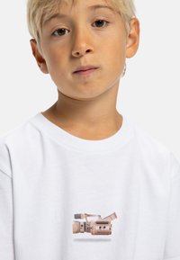 Camiseta blanca de manga corta con un diseño impreso de una cámara de video vintage en suave beige y gris, con detalles en negro, situada en el centro del pecho.